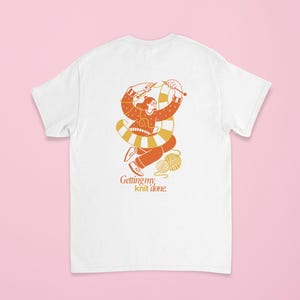 May include: White t-shirt featuring an orange and yellow illustration of a person knitting. The illustration includes a ball of yarn and the text "Getting my knit done."
