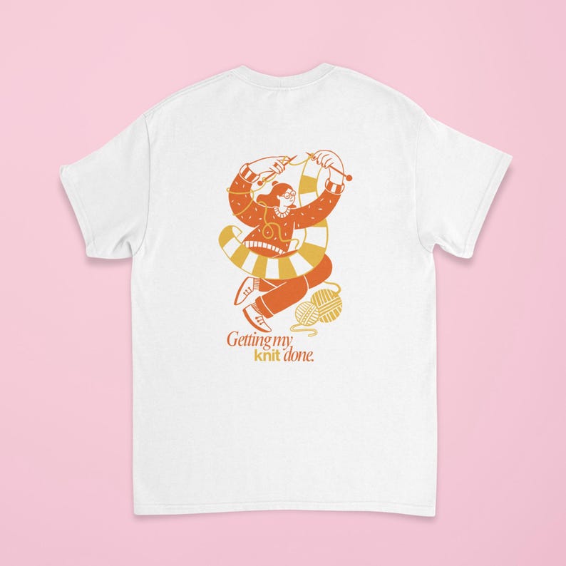 May include: White t-shirt featuring an orange and yellow illustration of a person knitting. The illustration includes a ball of yarn and the text "Getting my knit done."