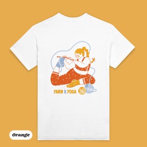 Op de afbeelding: Wit T-shirt met een afbeelding van een persoon die yoga doet en breit. De illustratie is in oranje, blauw en geel, met de woorden "Yarn and Yoga" eronder. Het T-shirt heeft een ronde hals.