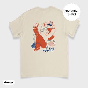 May include: Cream-colored t-shirt with an illustration of a person knitting, with orange clothing and blue yarn balls. The text "Knit happens" is below the illustration. The shirt also has the text "Natural Shirt" on it.