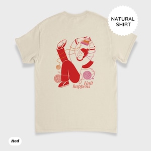 May include: Cream-colored t-shirt with a red graphic of a person knitting, surrounded by yarn balls. The text "Knit happens" is below the graphic. The shirt has a "Natural Shirt" label and a "Red" label.