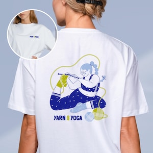 Op de afbeelding: Wit T-shirt met een blauw en geel ontwerp van een persoon die breit in een yoga pose. De tekst "YARN & YOGA" staat op de voor- en achterkant. Het shirt heeft een ronde hals en korte mouwen.
