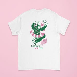 May include: White t-shirt with a graphic of a person knitting, wearing glasses and a green sweater. The person is surrounded by a pink and white striped design and green yarn. The text "Getting my knit done" is below the graphic.