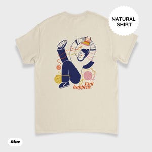 May include: Cream-colored t-shirt with a graphic of a person knitting, surrounded by yarn balls. The text "Knit happens" is below the illustration. The shirt also has the text "Natural Shirt" and "Blue" printed on it.