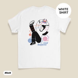 May include: White t-shirt featuring a whimsical illustration of a person knitting with colorful yarn balls. The design includes the text "Knit happens" in a playful font. The shirt has a round neck and short sleeves.