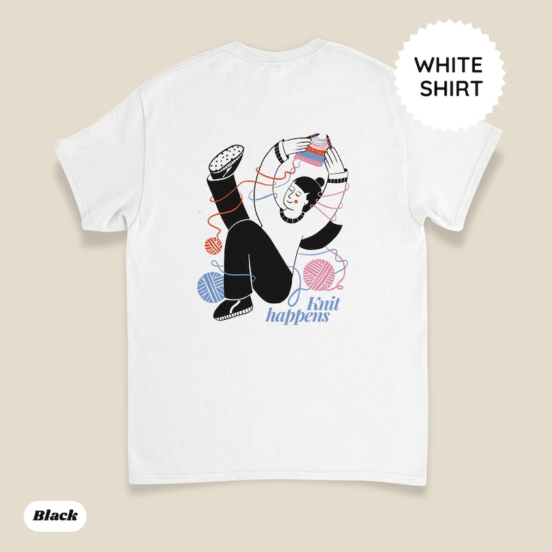 May include: White t-shirt featuring a whimsical illustration of a person knitting with colorful yarn balls. The design includes the text "Knit happens" in a playful font. The shirt has a round neck and short sleeves.