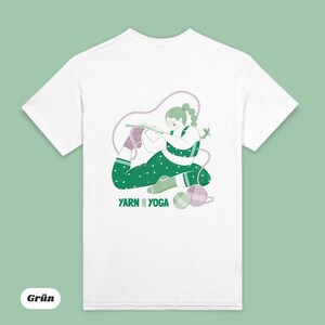 Op de afbeelding: Wit t-shirt met een groen en paarse illustratie van een persoon die in een yogahouding breit. De persoon draagt een groene legging met stippen en een bijpassende top. De tekst "YARN YOGA" staat eronder.