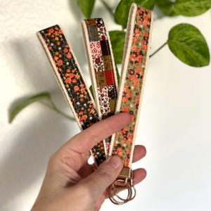 May include: Three floral wrist keychains with gold-colored hardware. The keychains feature a variety of patterns, including a dark floral print, a patchwork design, and a floral print on a tan background. Each keychain has a cream-colored border.