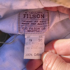 May include: Close-up of a FILSON label with the text "A GENUINE FILSON" and "MIGHT AS WELL HAVE THE BEST." The label also includes "C.C. FILSON CO. SEATTLE," lot number 79, and size 30. The bottom of the label reads "100% Cotton."