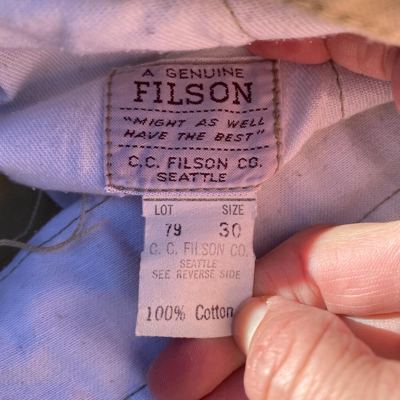 May include: Close-up of a FILSON label with the text "A GENUINE FILSON" and "MIGHT AS WELL HAVE THE BEST." The label also includes "C.C. FILSON CO. SEATTLE," lot number 79, and size 30. The bottom of the label reads "100% Cotton."