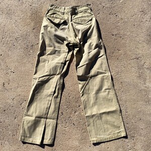 May include: Khaki-colored pants with a wide leg design. The pants have two back pockets and a button and zipper closure. The fabric appears to be a sturdy cotton or blend. The pants are laid out on a textured surface.