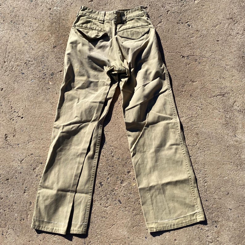 May include: Khaki-colored pants with a wide leg design. The pants have two back pockets and a button and zipper closure. The fabric appears to be a sturdy cotton or blend. The pants are laid out on a textured surface.