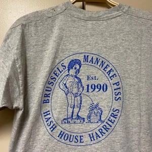 Puede incluir: Camiseta gris con un gráfico circular azul de una estatua de un niño orinando. El gráfico incluye el texto "Manneke Pis Harriers Hash House Brussels Est. 1990".