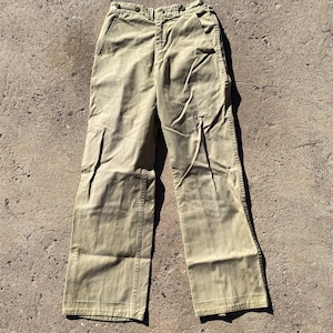 May include: Khaki-colored pants with a classic design. The pants have a button closure and pockets. The fabric appears to be a sturdy cotton or blend. The pants are laid flat on a concrete surface.