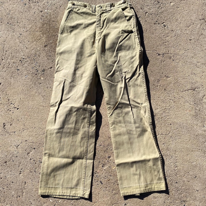 May include: Khaki-colored pants with a classic design. The pants have a button closure and pockets. The fabric appears to be a sturdy cotton or blend. The pants are laid flat on a concrete surface.