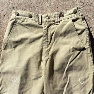 May include: Close-up of khaki-colored pants. The pants have a button and zipper closure, belt loops, and pockets. The fabric appears to be a sturdy cotton or blend. The pants have a classic, utilitarian design.