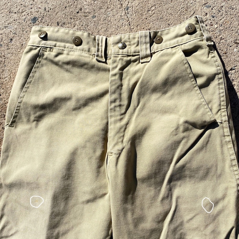 May include: Close-up of khaki-colored pants. The pants have a button and zipper closure, belt loops, and pockets. The fabric appears to be a sturdy cotton or blend. The pants have a classic, utilitarian design.
