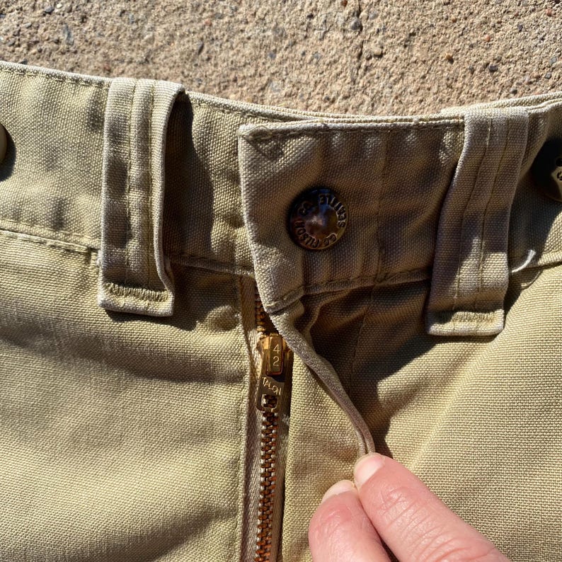 May include: Close-up of khaki-colored pants with a brass zipper and button closure. The zipper is partially open, revealing the Talon brand. The pants have belt loops and a button with the text "FILSON" on it.