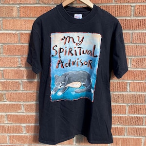 May include: Black t-shirt with a colorful graphic featuring the text "My Spiritual Advisor" above a gray cat illustration. The cat is lying down on a blue background. The shirt is hanging on a wooden hanger.