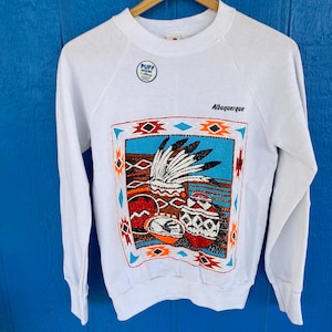 May include: White crew neck sweatshirt with a colorful Southwestern design. The graphic features Native American pottery and a headdress, with the word "Albuquerque" printed above the design. A small "Puff Design" logo is on the upper left.