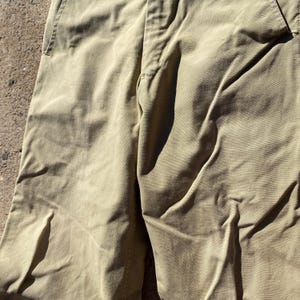 May include: Close-up of khaki-colored pants. The pants have a button and zipper closure, belt loops, and pockets. The fabric has visible texture and folds, suggesting a casual style. The color is a muted, earthy tone.