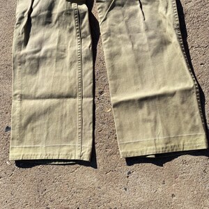 May include: Close-up of khaki-colored pants. The pants have a wide leg design and are made of a durable fabric. The stitching is visible along the seams and the bottom hem. The pants are folded and lying on a rough surface.