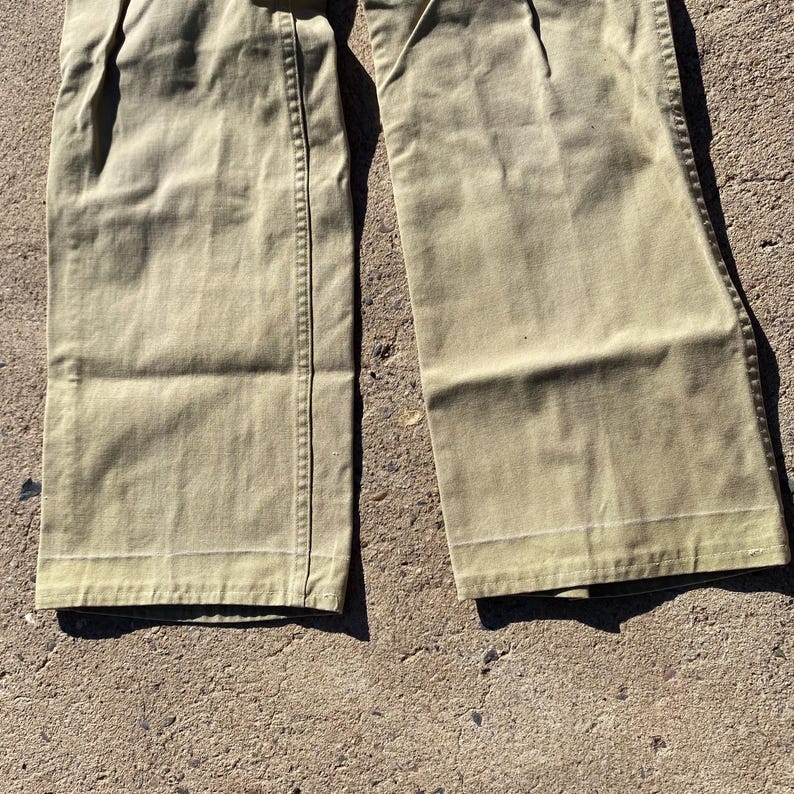 May include: Close-up of khaki-colored pants. The pants have a wide leg design and are made of a durable fabric. The stitching is visible along the seams and the bottom hem. The pants are folded and lying on a rough surface.