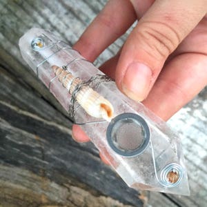 May include: A clear quartz crystal pipe with a seashell and wire wrapped around it. The pipe has a metal screen and a small hole for airflow.