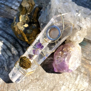 May include: A clear quartz crystal pipe with a metal bowl and amethyst crystals wrapped around it with gold wire. The pipe is sitting on a wooden surface with other crystals.