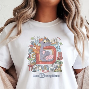 May include: White t-shirt featuring a colorful graphic of a cartoon dragon inside a letter "D" with the text "Walt Disney World" below. The design includes various park elements and is in a vintage style.