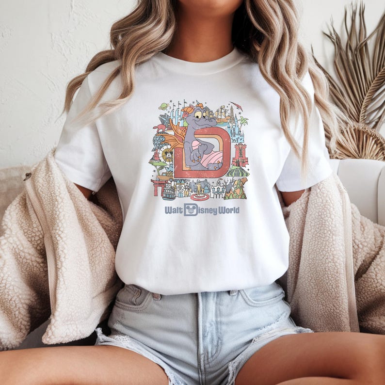 May include: White t-shirt featuring a colorful graphic of a cartoon dragon inside a letter "D" with the text "Walt Disney World" below. The design includes various park elements and is in a vintage style.