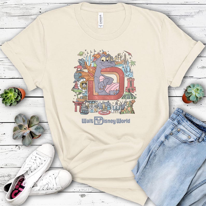 May include: Cream-colored t-shirt featuring a colorful graphic of a purple dragon inside a red letter "D" with "Walt Disney World" text. The design includes various Disney-themed elements. The shirt is paired with blue jeans and white sneakers.