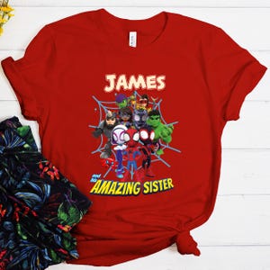 Puede incluir: Camiseta roja con un dise&ntilde;o de superh&eacute;roes de dibujos animados, incluyendo a Spider-Man y Hulk, con el nombre "JAMES" arriba y "and his AMAZING SISTER" abajo. El dise&ntilde;o est&aacute; sobre una telara&ntilde;a azul.
