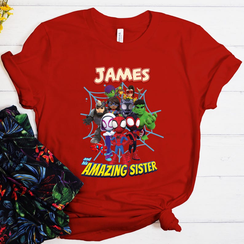 Puede incluir: Camiseta roja con un dise&ntilde;o de superh&eacute;roes de dibujos animados, incluyendo a Spider-Man y Hulk, con el nombre "JAMES" arriba y "and his AMAZING SISTER" abajo. El dise&ntilde;o est&aacute; sobre una telara&ntilde;a azul.