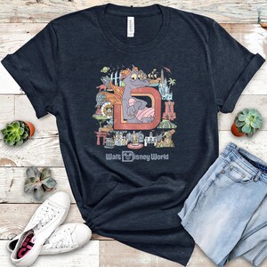 May include: Navy blue t-shirt featuring a colorful graphic of a dragon character inside a letter "D" with various Disney World landmarks. The text "Walt Disney World" is printed below the graphic. The shirt is paired with blue jeans and white sneakers.