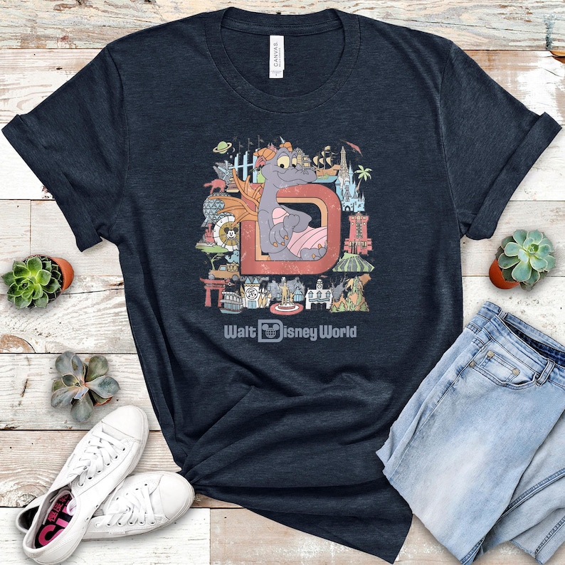May include: Navy blue t-shirt featuring a colorful graphic of a dragon character inside a letter "D" with various Disney World landmarks. The text "Walt Disney World" is printed below the graphic. The shirt is paired with blue jeans and white sneakers.