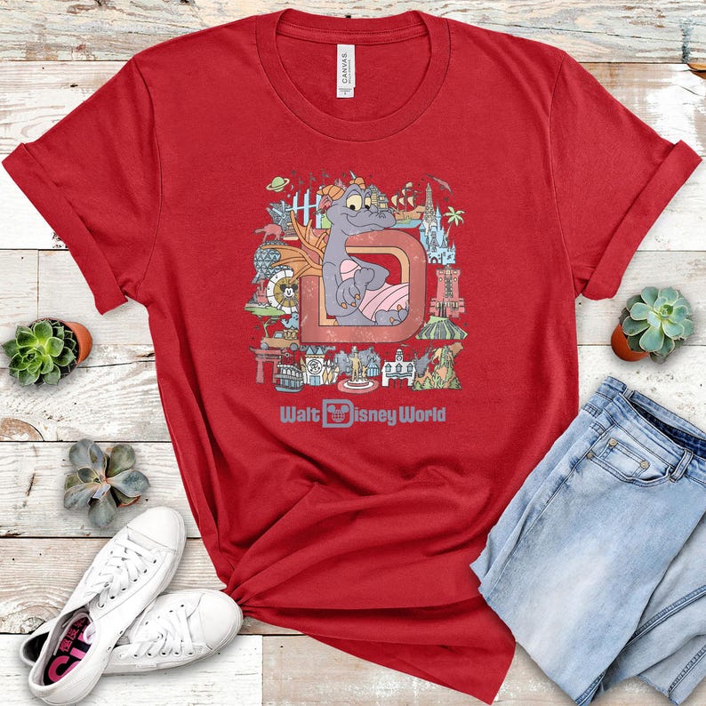 May include: Red t-shirt featuring a colorful graphic of a purple dragon character, the letter "D", and various Walt Disney World landmarks. The text "Walt Disney World" is printed below the graphic. The shirt is paired with blue jeans and white sneakers.