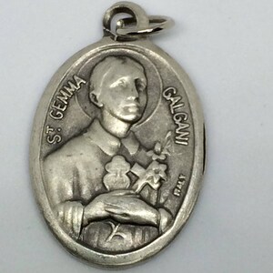 Può includere: Un ciondolo ovale in metallo color argento con un ritratto in rilievo di Santa Gemma Galgani che tiene in mano una piccola croce e un cuore. Il testo ST. GEMMA GALGANI è impresso sui lati, con la parola ITALY vicino al bordo inferiore.