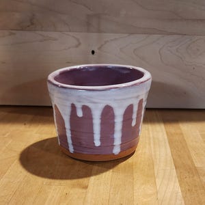 May include: A handmade ceramic bowl with a maroon exterior and a white, dripped glaze design. The interior is a deep purple colour. The bowl sits on a natural, unglazed base. The bowl is on a wooden surface.