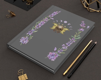 May include: Gray hardcover journal with a floral design featuring purple flowers and a skull with wings. The skull has a star and crescent moon design.