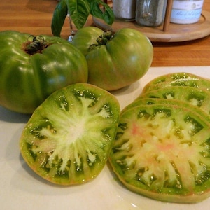 Può includere: Pomodori verdi tagliati a fette per rivelare un interno verde chiaro con un centro rosa. I pomodori sono su una superficie bianca.