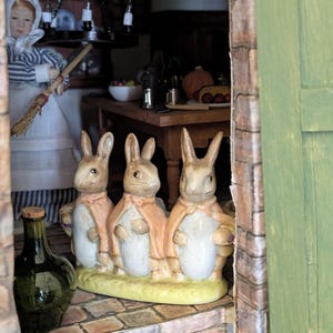 Puede incluir: Escena en miniatura con tres figuras de conejos con capas rosas sobre una base verde. Se ve una botella verde con tapón de corcho. La escena está ambientada en una cocina en miniatura.