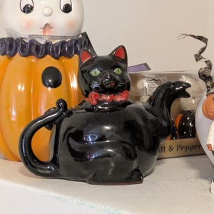 Op de afbeelding: Een zwarte katvormige theepot met rode oren en een rode strik. De theepot heeft groene ogen en een glanzende afwerking. Andere Halloween-decoraties zijn zichtbaar op de achtergrond.