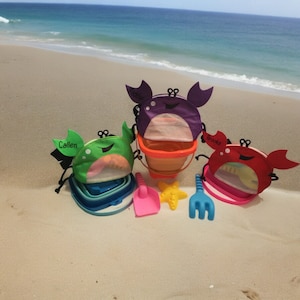 May include: Three colorful crab-shaped beach pails with mesh bottoms and personalized names: "Cullen", "Ryder", and "Brooks". The pails are on a sandy beach with a blue ocean in the background. There are also a pink shovel and a blue rake.