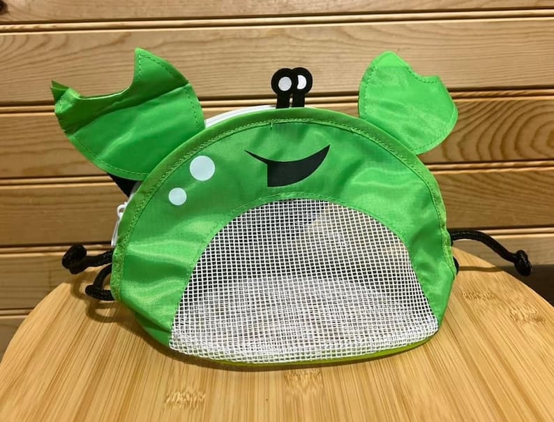 May include: Green mesh drawstring bag with a crab design. The bag has a white mesh bottom and a green fabric top with a black smiling face and white eyes.