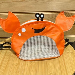 May include: Orange and black crab-shaped mesh bag with a drawstring closure. The bag has a smiling face with two eyes and a black mouth.