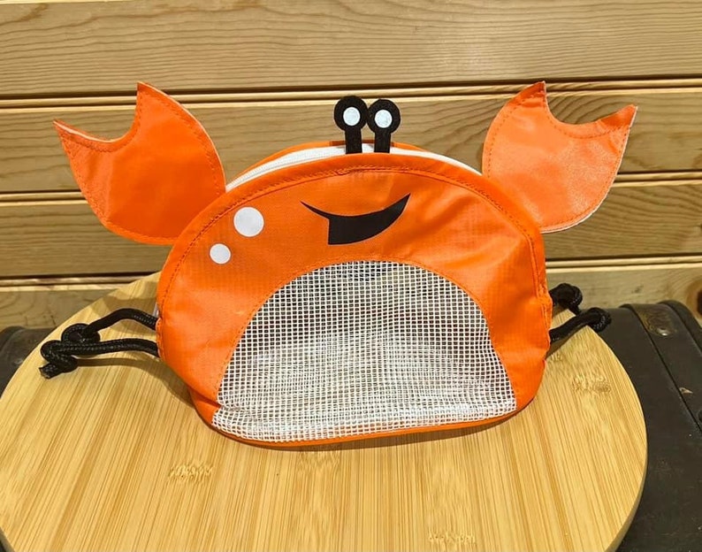 May include: Orange and black crab-shaped mesh bag with a drawstring closure. The bag has a smiling face with two eyes and a black mouth.