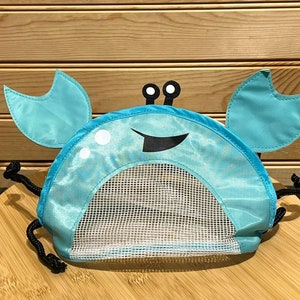 May include: A blue crab-shaped mesh toy bag with black drawstring ties. The bag has white eyes and a black smiling mouth.