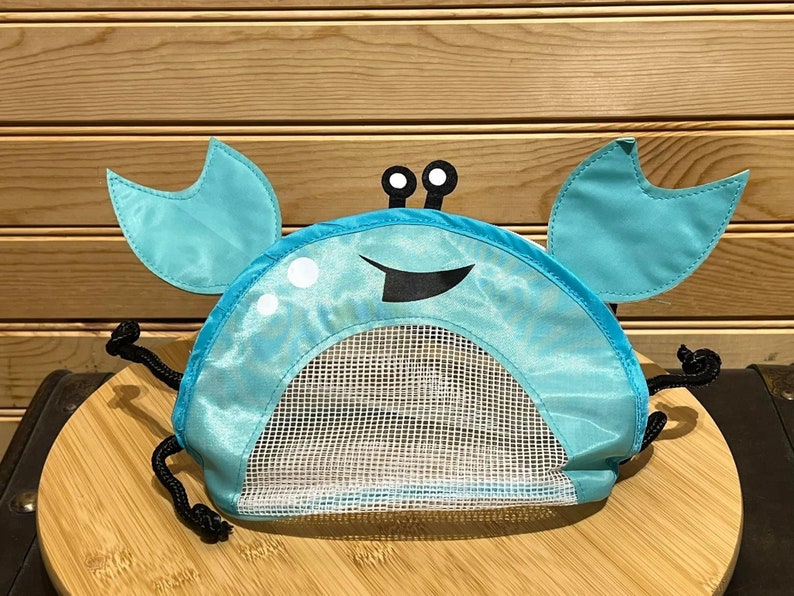 May include: A blue crab-shaped mesh toy bag with black drawstring ties. The bag has white eyes and a black smiling mouth.
