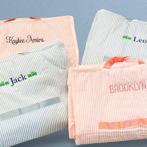 May include: Four personalized garment bags with striped fabric. The bags are in two colors: pink and blue. The names "Kaylee Amira", "Jack", "Leo", and "Brooklyn" are embroidered on the bags.